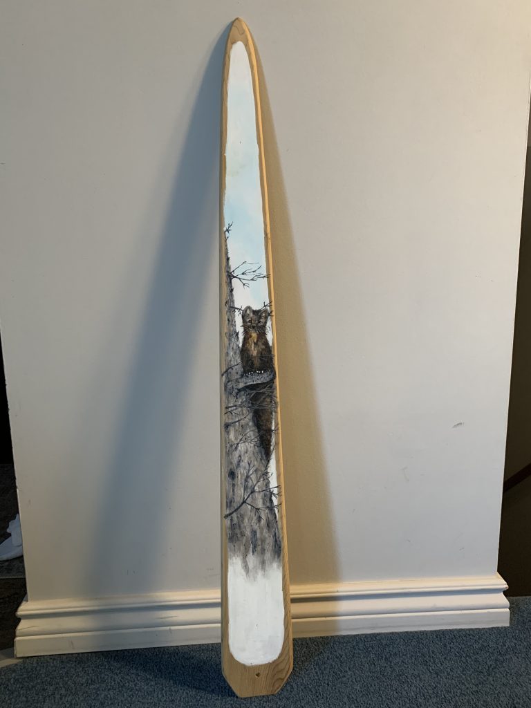Tall wooden paddle with a detailed painting of a lynx perched on a tree branch against a pale blue sky background.