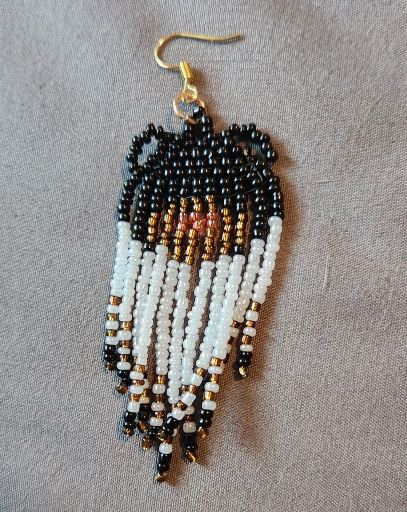 Handcrafted earring made of black, white, and gold beads arranged in a fringe pattern with a gold hook on a gray fabric background.