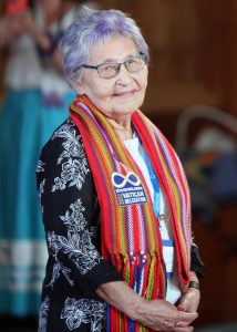 Elder Angie Mercredi-Crerar smiles while wearing a sash and clasping her hands. She's wearing a black and white floral blouse and her hair is slightly purple and while. She is wearing black glasses